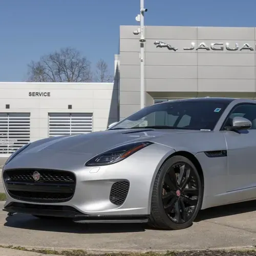 A silver Jaguar F-TYPE sports car parked in front of a Jaguar dealership on a sunny day.