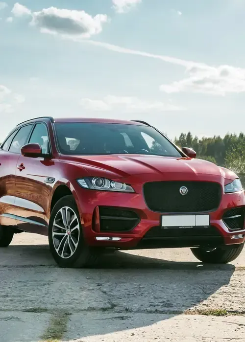 A red Jaguar SUV parked on a paved lot under a sunny, cloudy sky.