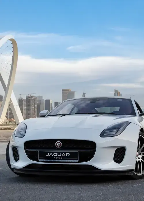 A white Jaguar F-Type sports car parked in front of a city skyline with a large, white abstract sculpture nearby.