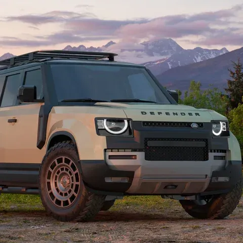 A tan Land Rover Defender SUV with a roof rack parked on a dirt path in front of mountains at sunset.