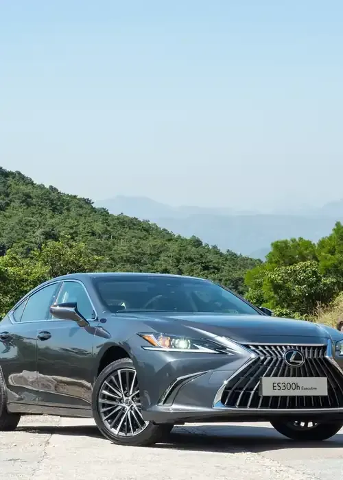 A dark grey Lexus ES sedan parked on a road with a scenic, green, mountainous background under a clear sky.