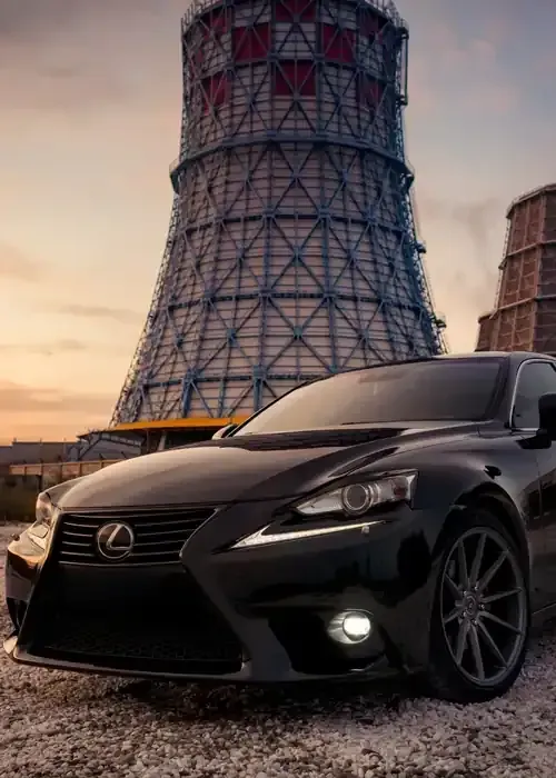 A sleek black Lexus sedan parked on gravel in front of a large industrial cooling tower at sunset.