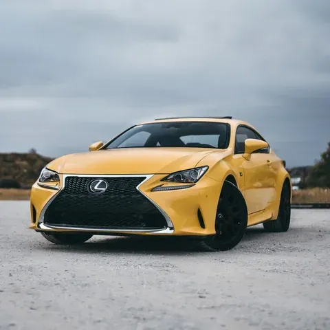A bright yellow Lexus coupe parked on a gravel surface under a cloudy sky.