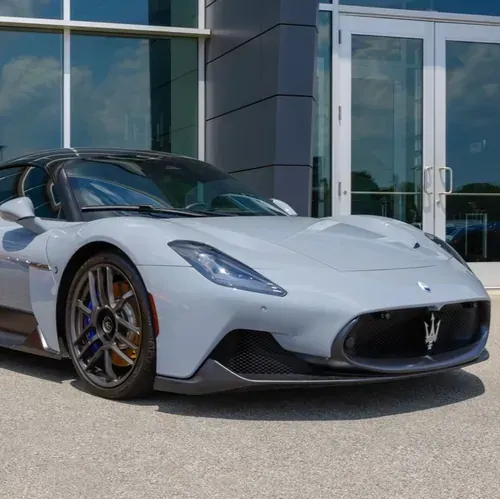 A gray Maserati MC20 sports car parked in front of a glass-fronted building.
