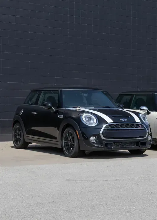 A black MINI Cooper with white hood stripes parked on asphalt in front of a dark gray wall.