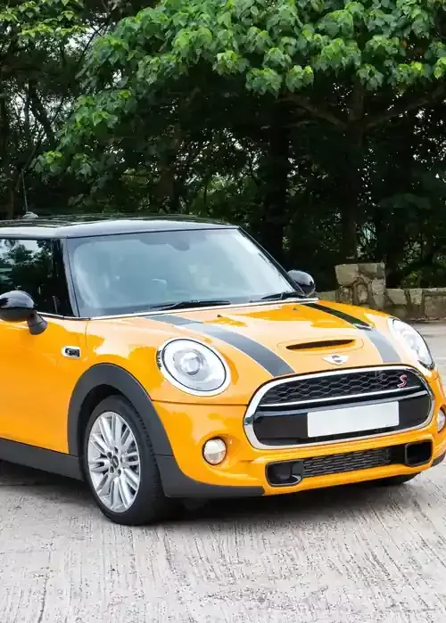 A bright yellow Mini Cooper S with black hood stripes parked on a gravel surface in front of green trees.
