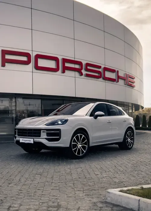 A white Porsche Cayenne SUV parked in front of a modern dealership building with large red Porsche lettering.