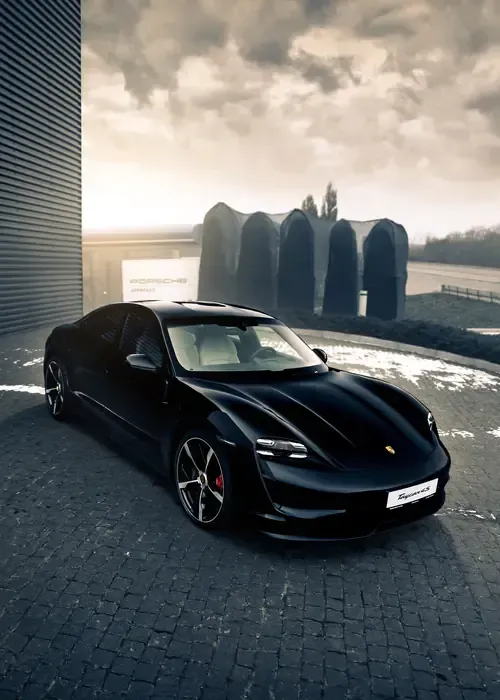 A black Porsche Taycan parked on a paved driveway in front of a modern building with arched architectural features.