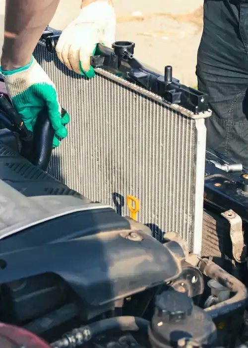 Mechanic in green and tan gloves holding an engine radiator over an open car hood.