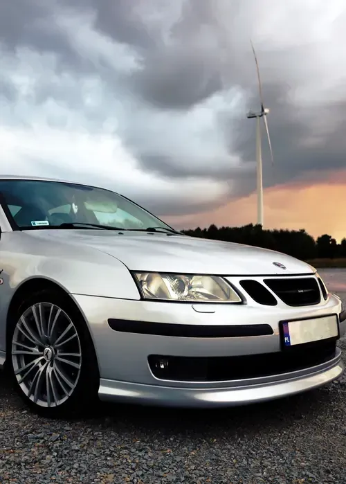 A silver Saab sedan parked on a gravel surface under a dramatic, cloudy sky with a wind turbine in the background.