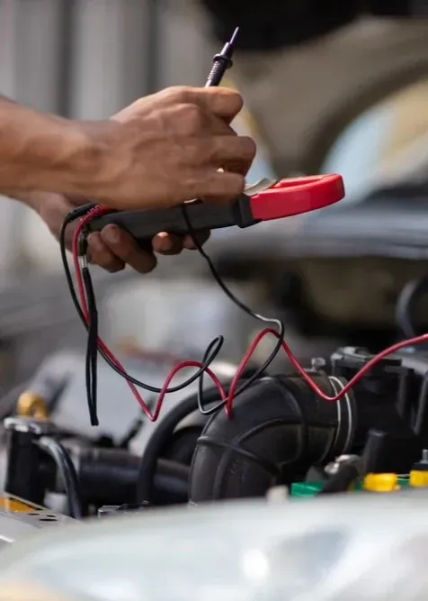 A person holding a red and black digital multimeter to test an engine under the hood of a car.