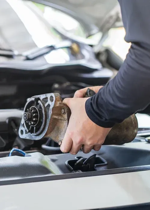 A mechanic holding a used car starter motor over an open vehicle engine bay.