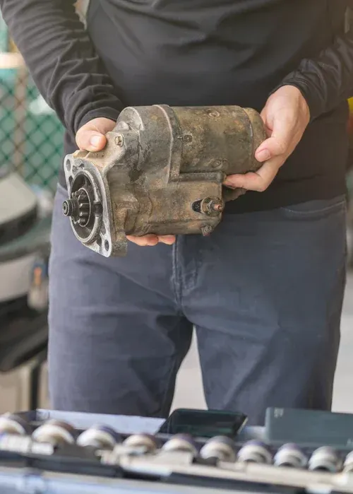 A person in a long-sleeved shirt holds a dirty automotive starter motor in their hands.