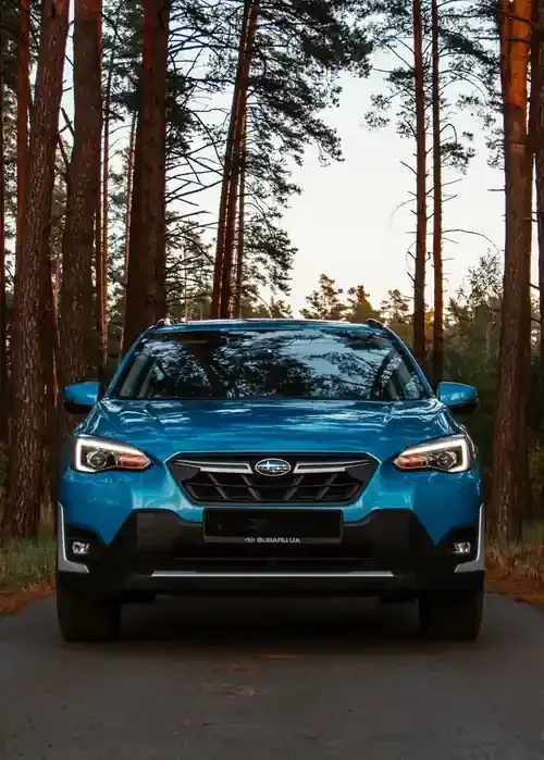 A bright blue Subaru Crosstrek parked on a paved road surrounded by a forest of tall pine trees.