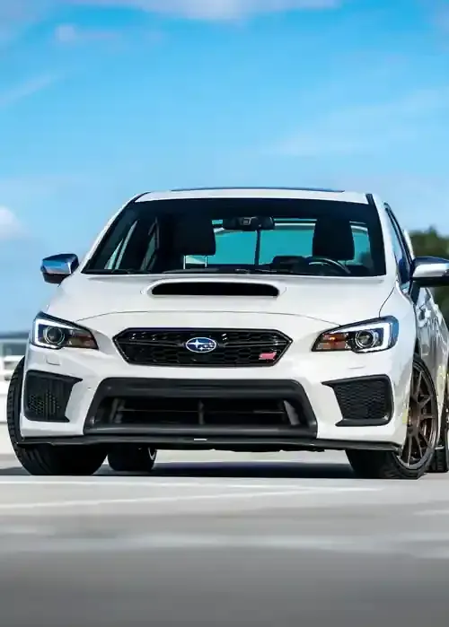 A white Subaru WRX STI parked on a paved surface under a bright blue sky.