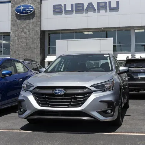 A silver Subaru Legacy parked in front of a Subaru dealership under a bright, sunny sky.