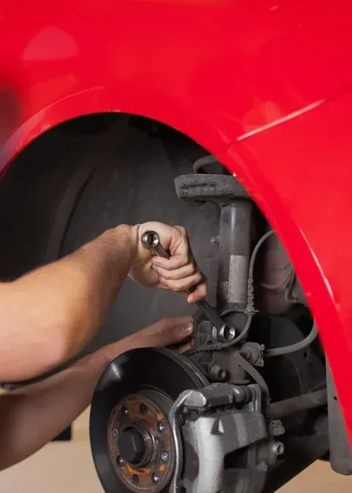 A mechanic uses a wrench to work on the suspension system of a red car inside an auto repair shop.