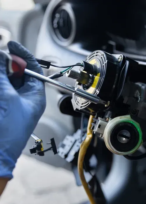 A gloved hand uses a screwdriver to work on the clock spring behind a removed car steering wheel.