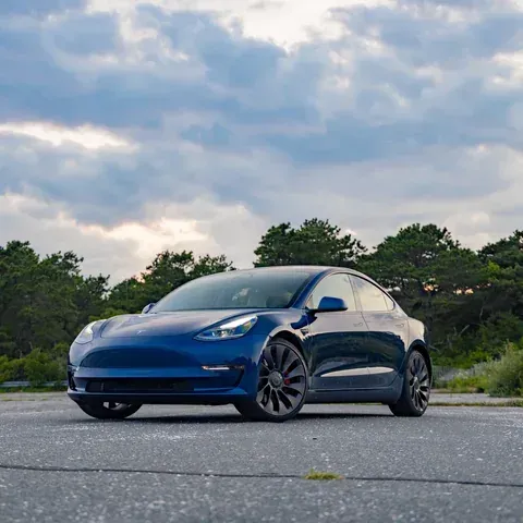 A deep blue Tesla Model 3 parked on an asphalt surface against a background of green trees and a cloudy sky.