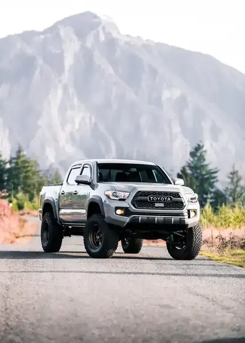 A silver Toyota Tacoma truck driving on a paved road with a large, misty mountain in the background.
