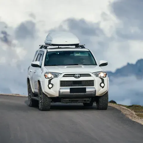 A white Toyota 4Runner with a roof cargo box driving on a winding paved mountain road under a cloudy sky.