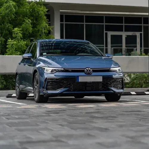 A blue Volkswagen Golf parked on a paved lot in front of a modern building with glass windows.