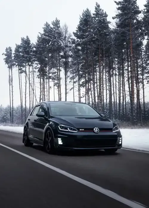 A dark Volkswagen Golf GTI driving on an asphalt road bordered by snow-covered pine trees in a winter landscape.