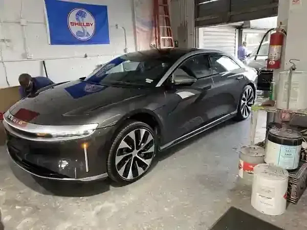 A dark grey Lucid Air sedan parked inside a garage with a Shelby flag on the wall and supplies nearby.