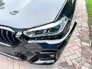 Close-up of a black car’s front headlight and grille on a brick driveway after rain