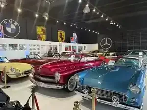 Classic cars displayed in a museum, including a red convertible and blue roadster under hanging lights
