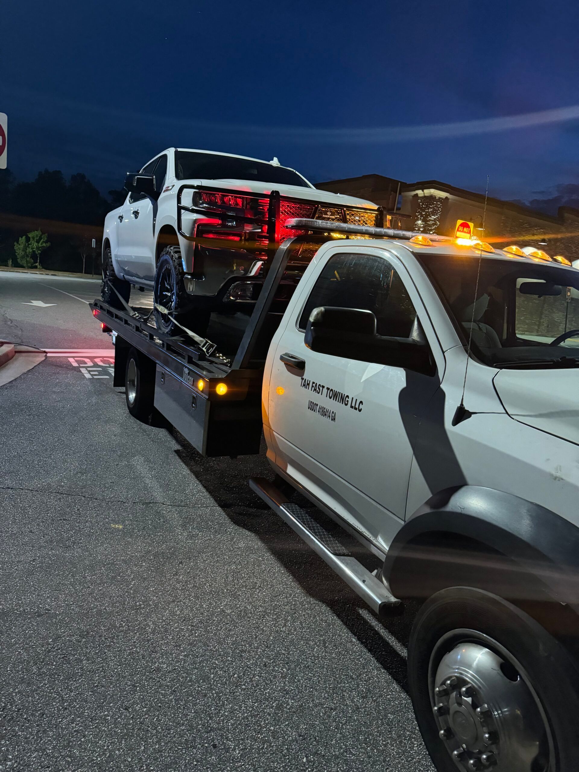 A tow truck is towing a white truck in a parking lot at night.