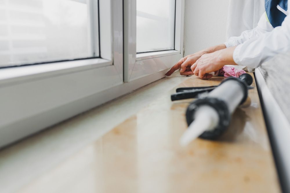 Hands Applying Caulk Along a White Window Frame Beside a Tube of Sealant — ACG Windows & Glass in Cheltenham, VIC