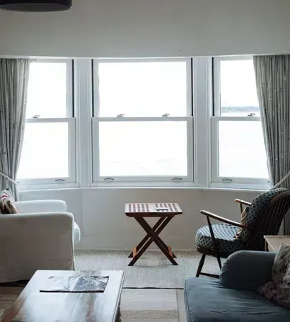 Bright living room with bay windows, gray curtains, wicker chair, sofa, and small wooden side table — ACG Windows & Glass in Frankston, VIC