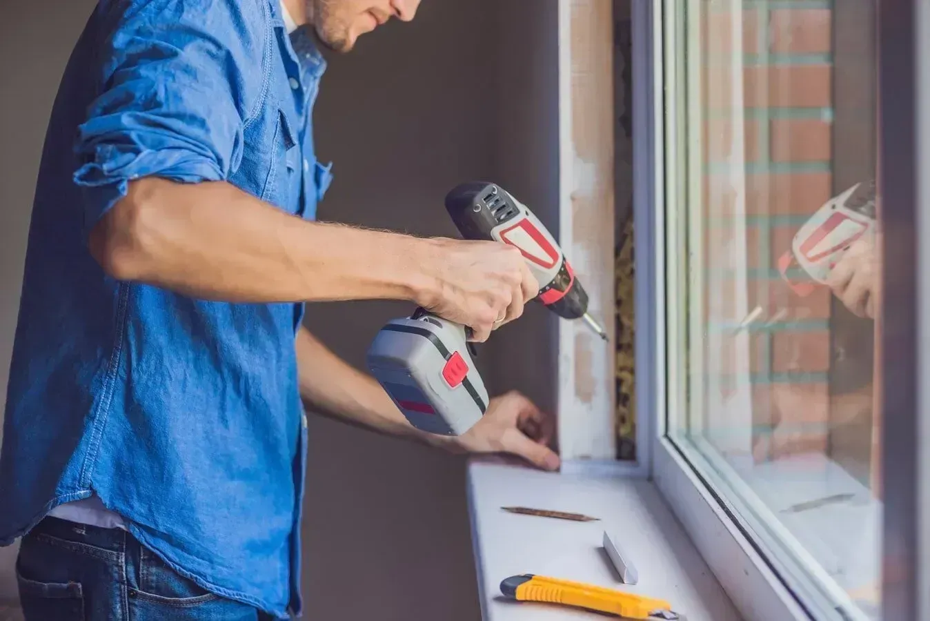 A Person in a Blue Shirt Uses a Power Drill to Install a White Window — ACG Windows & Glass in Frankston, VIC