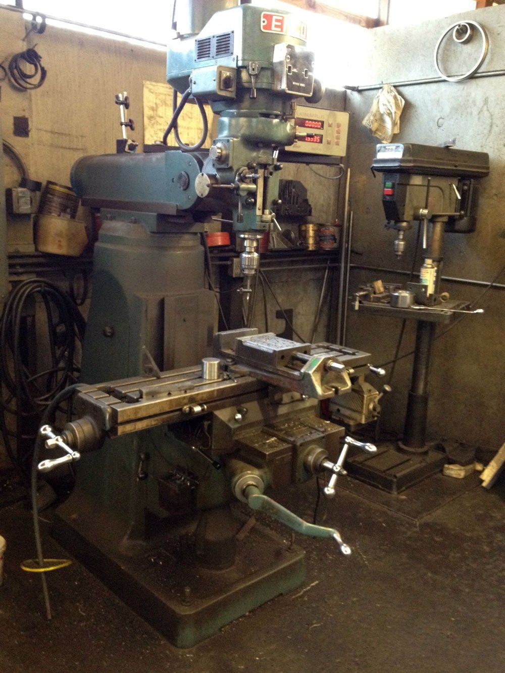 A milling machine in a workshop, with a drill press visible in the background.