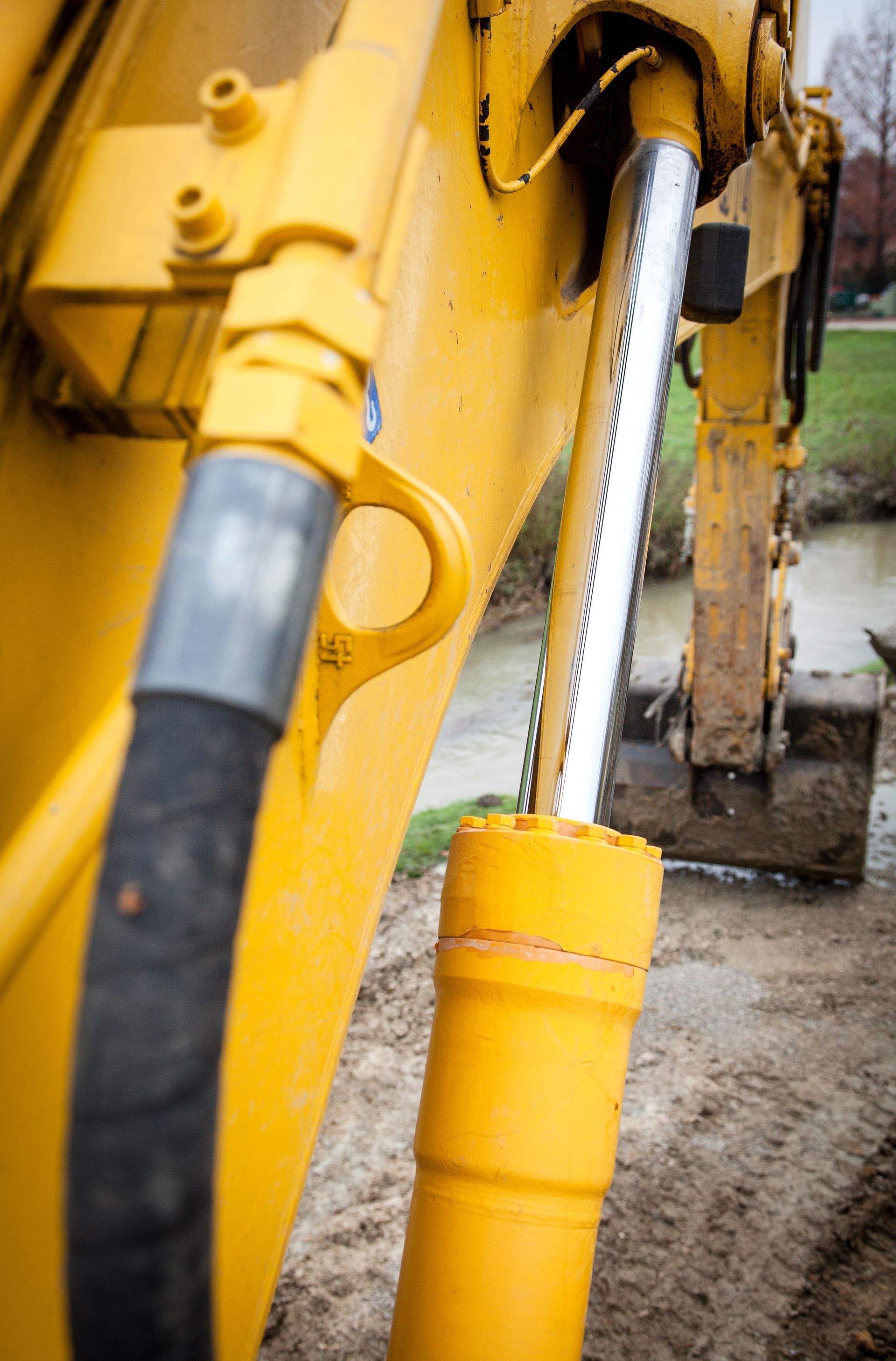 Yellow excavator arm with hydraulic cylinder, black hose, and dirt background.