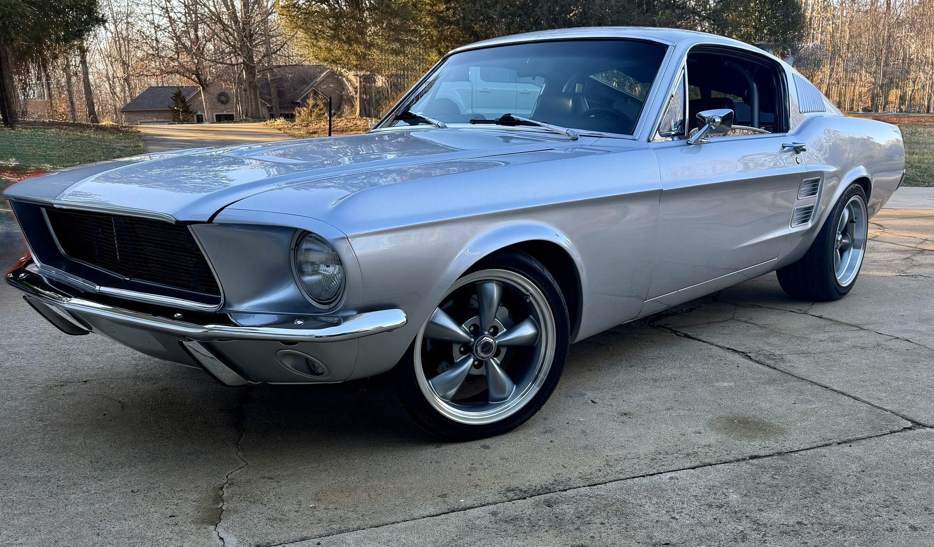 Silver 1967 Ford Mustang coupe on a driveway, chrome details, aftermarket wheels.