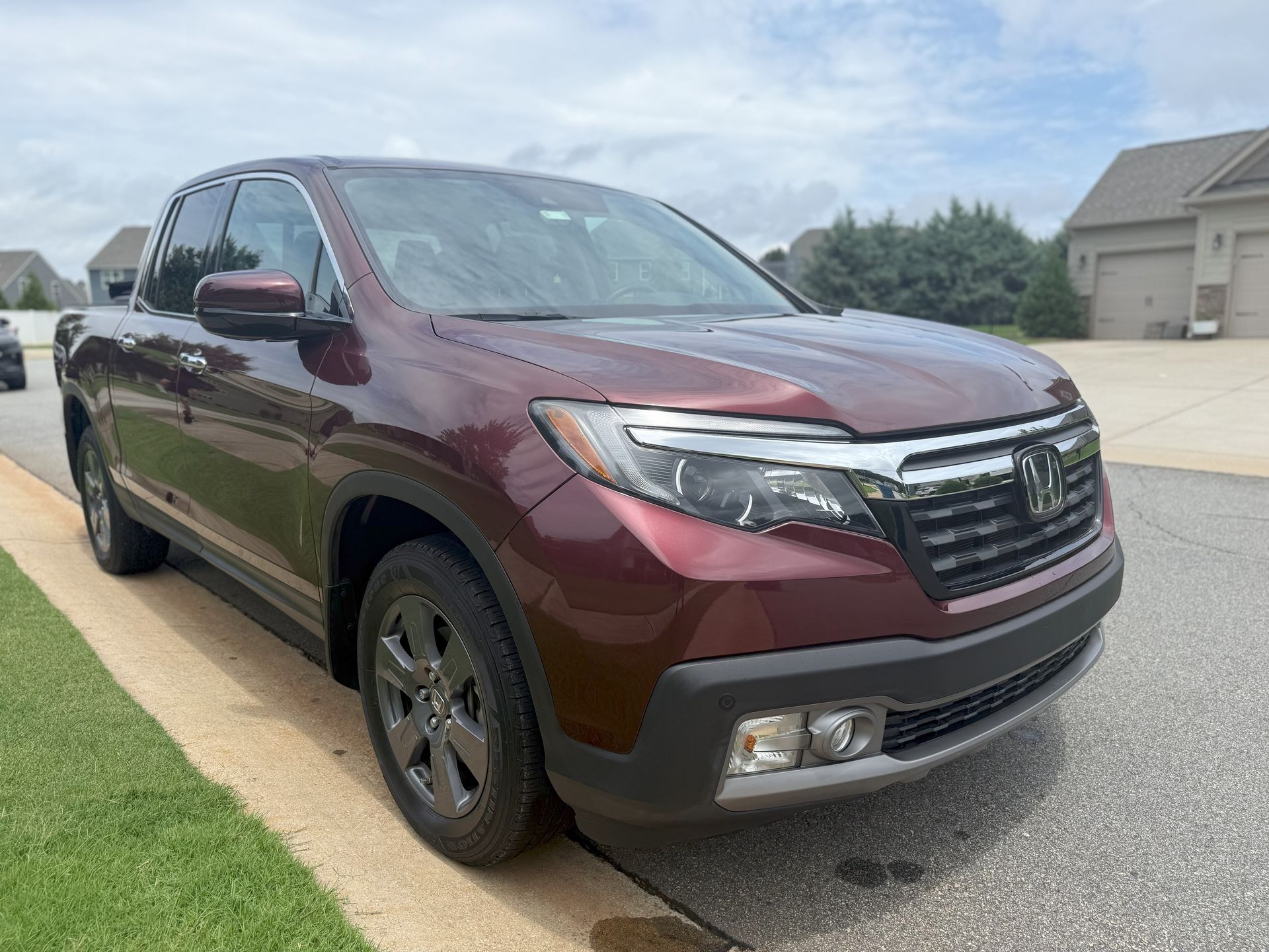 Maroon Honda Ridgeline pickup truck parked on a paved road next to a curb with green grass in front of houses.