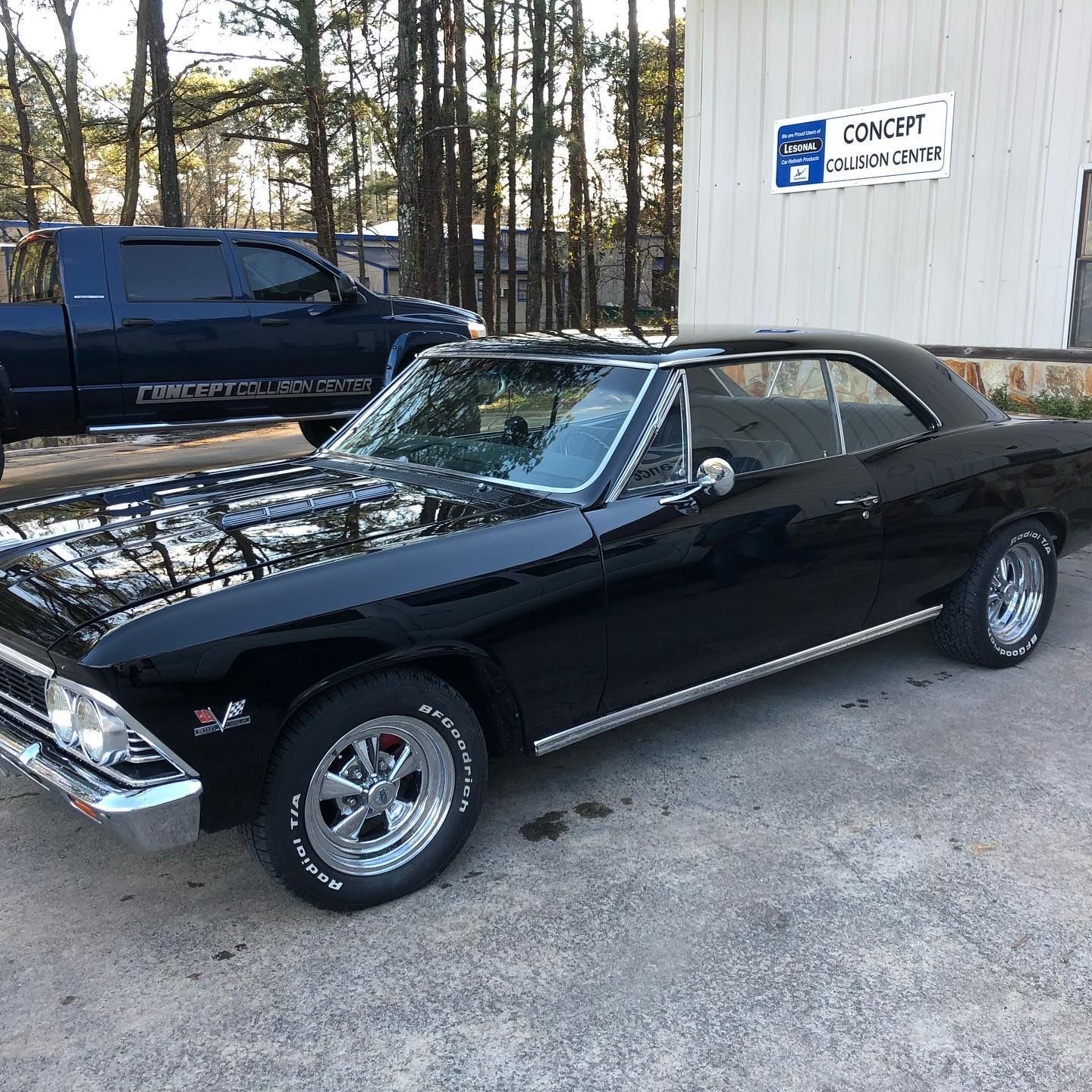 Black classic Chevrolet Chevelle parked outside of a building, with a dark blue truck in the background.