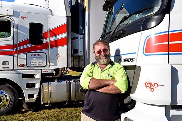 Man Standing With Arms Crossed, Smiling Next to Two White Trucks — Hines Logistic Pty Ltd In Forster, NSW