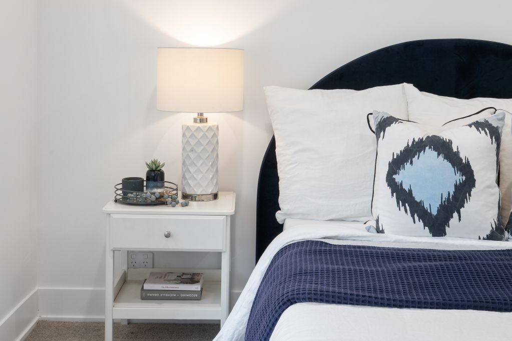 Cozy bedroom corner with a white nightstand lamp, blue bedding, and patterned accent pillow