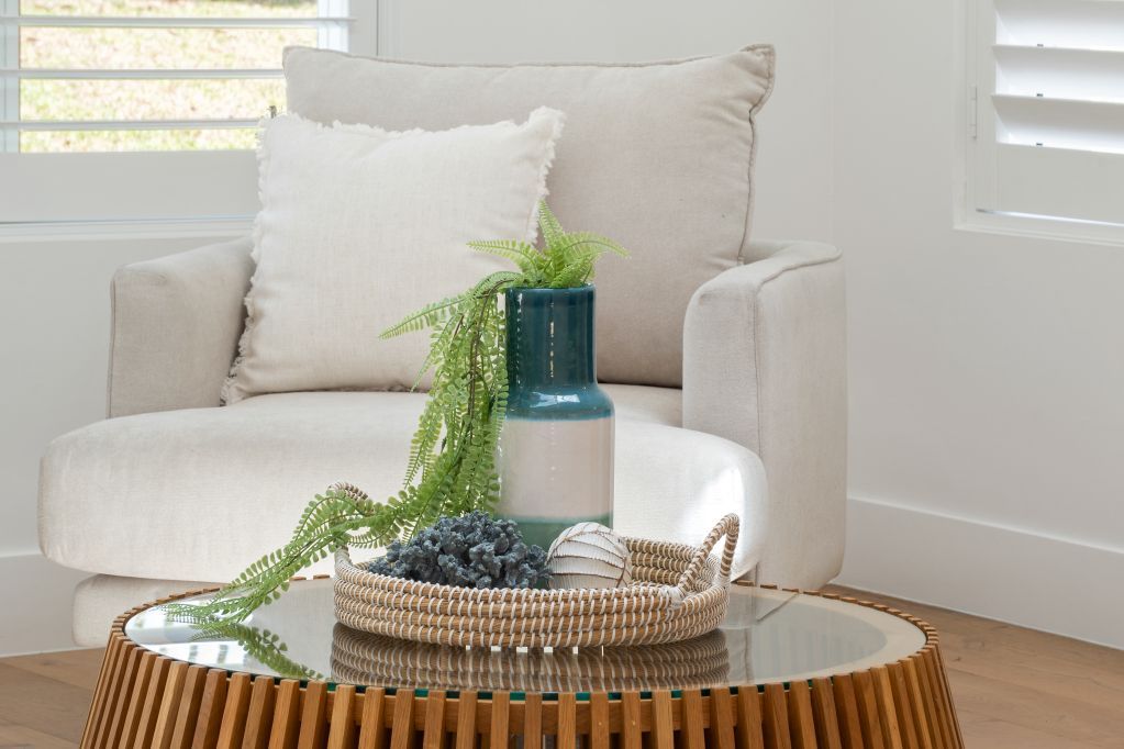 Cream upholstered chair beside a round coffee table with a woven tray, vase, and greenery in a bright room