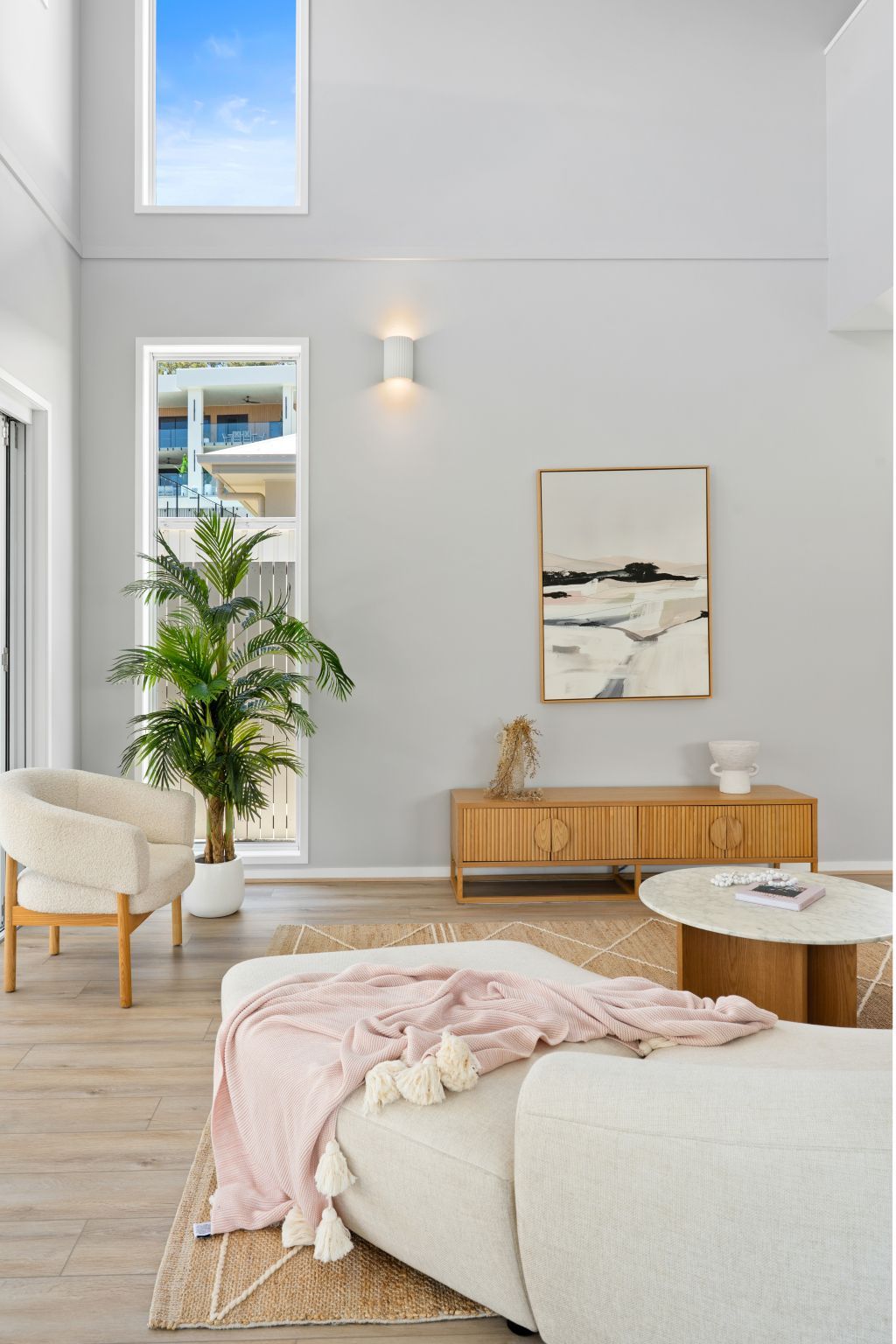Bright minimalist bedroom with white walls, a beige bed, wood console, plant, and soft pink throw blanket.