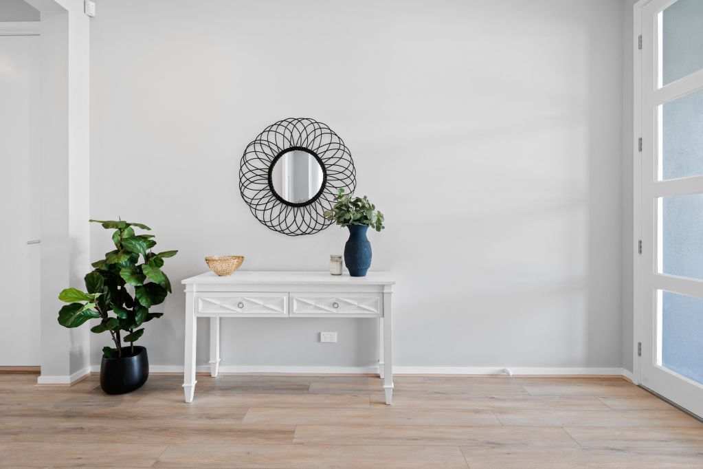 Bright minimalist room with white console table, round mirror, plants, and light wood floor near a blue door