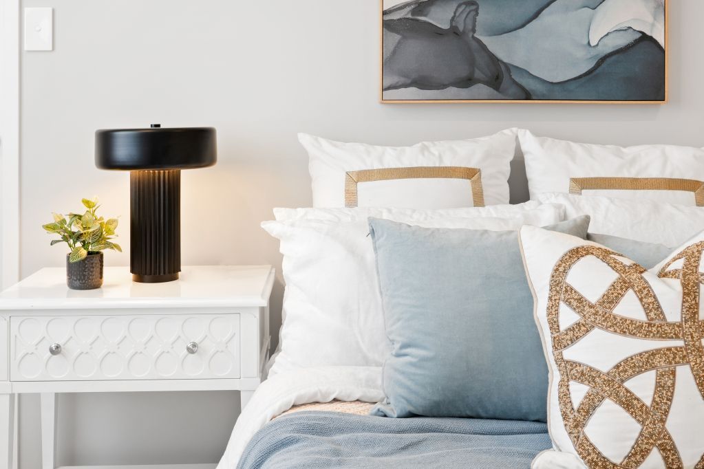 Bright bedroom with white bedding, pale blue accents, bedside lamp, and floral arrangement on a white nightstand