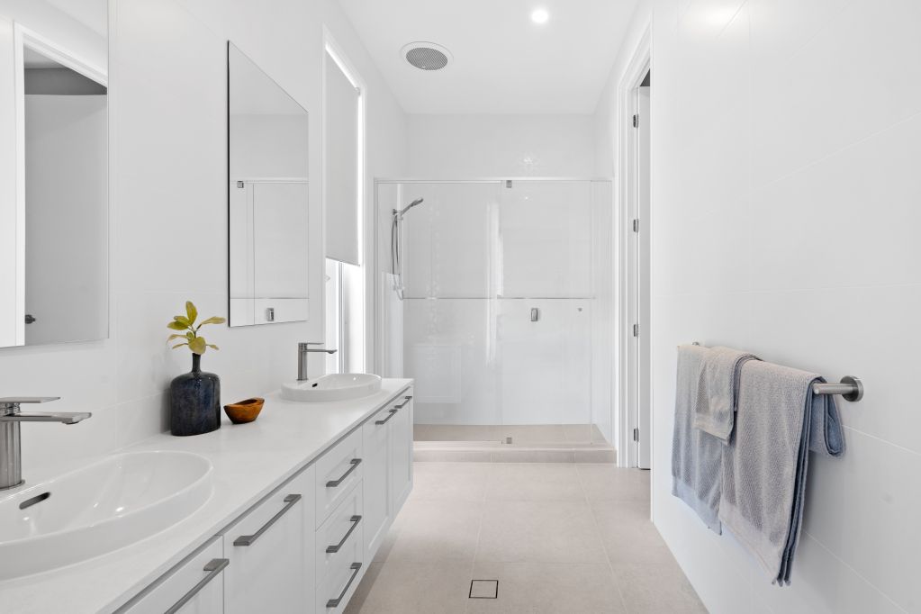 Bright white bathroom with double vanity, mirror, tub, and towels on a wall rack.
