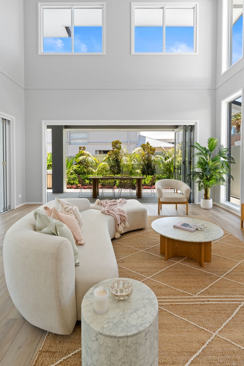 Bright modern living room with curved cream sofa, round table, woven rug, and open doors to a sunny patio.