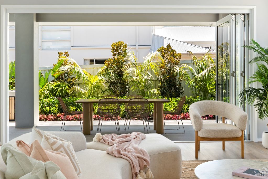 Bright living room opening to a lush garden, with white seating, pink throw, and wooden chairs.