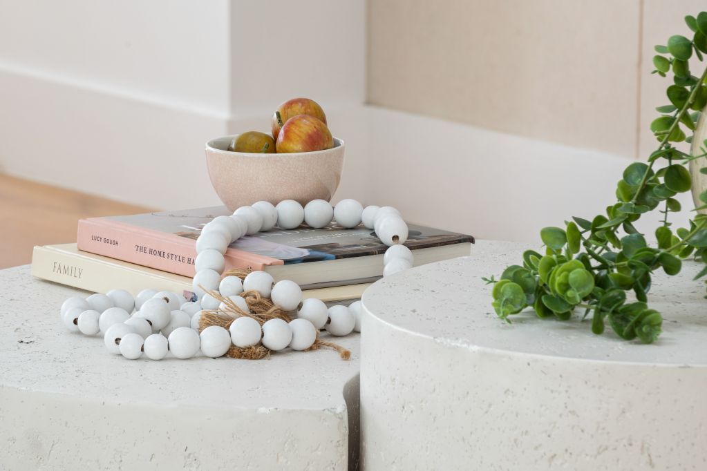 Minimalist tabletop display with stacked books, white beaded decor, fruit bowl, and green vine on a speckled surface