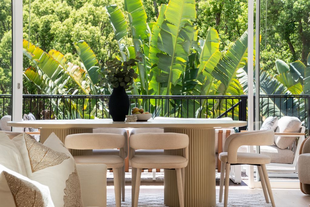 Bright patio with beige sofas, round table, and a vase of flowers overlooking lush green tropical plants.